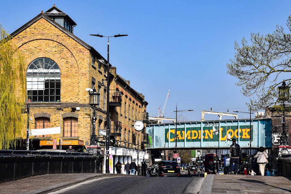 A busy street scene under a large, weathered railway bridge with the words 'Camden Lock' painted in large yellow letters on a blue-green metal sign attached to the bridge structure. The environment ap