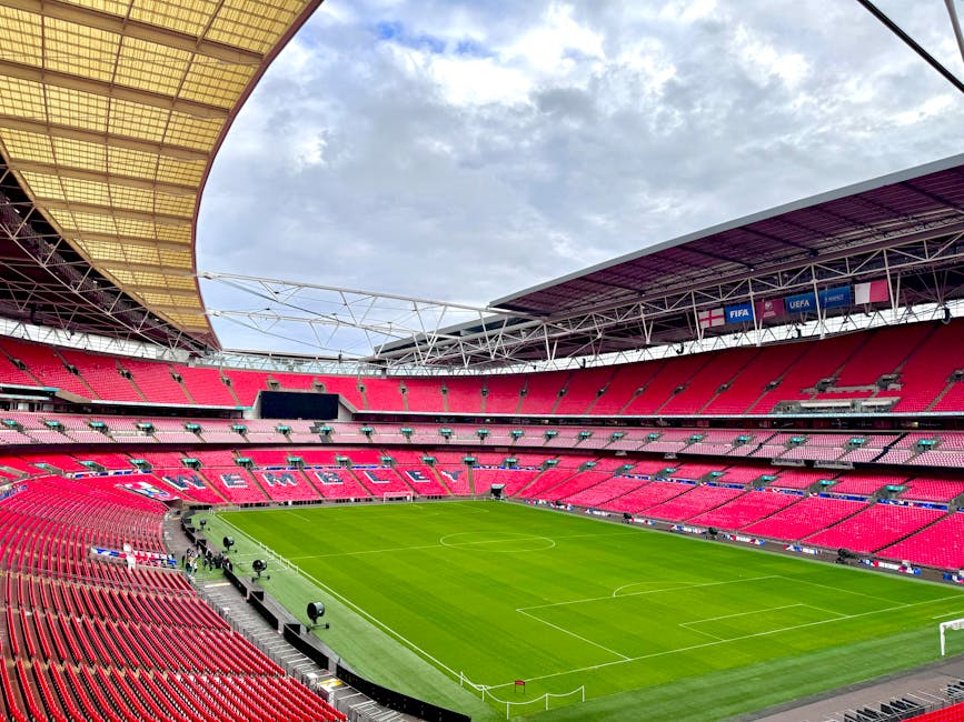 A large open-air football stadium with an extensive seating arrangement predominantly in red and pink tones surrounds a well-maintained green pitch. The seating sections are tiered, with some areas di